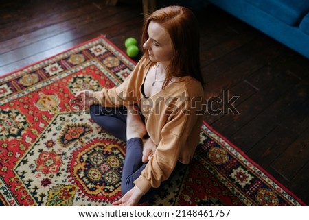A woman with red hair sits in a lotus position on the floor and meditates in her living room top view