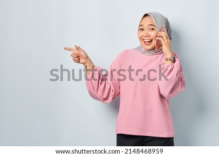 Cheerful young Asian woman in pink shirt talking on mobile phone and pointing finger away at blank space isolated over white background