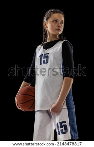 Portrait of teen girl, basketball player in uniform posing isolated over black studio background. Sport training. Basketball season. Concept of professional sport, childhood, motion, team game, ad