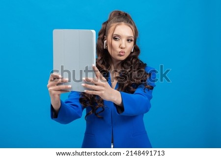 Portrait of a cheerful, positive, smiling woman holding a tablet, earphones in hands, taking a selfie against a blue background.