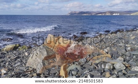The sea wave rolls over the coastal stones and foams up. One rock is very large. On the opposite shore of the bay, you can see mountains and settlements in the valleys. There is a cloudy sky Royalty-Free Stock Photo #2148501583
