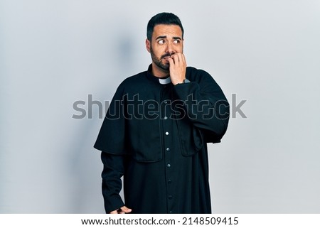 Handsome hispanic man with beard wearing catholic priest robe looking stressed and nervous with hands on mouth biting nails. anxiety problem. 
