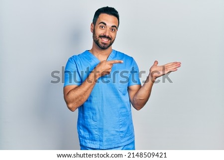 Handsome hispanic man with beard wearing blue male nurse uniform amazed and smiling to the camera while presenting with hand and pointing with finger. 