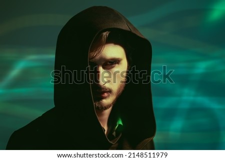 Studio portrait of a brutal brunette man looking intently and cautiously at the camera from under a black hood. Green background. People and emotions.  Royalty-Free Stock Photo #2148511979