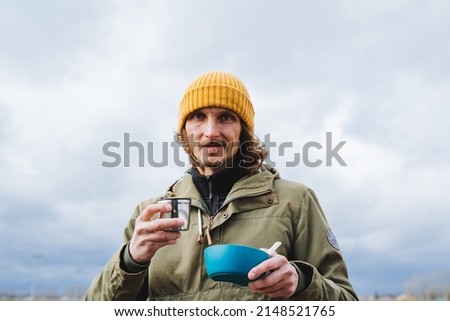Meals on a hike, a man holds in his hands a bowl of food and a mug of tea, lunch in the forest, a portrait against the sky, a tourist kitchen, a hipster on a walk there is soup. High quality photo
