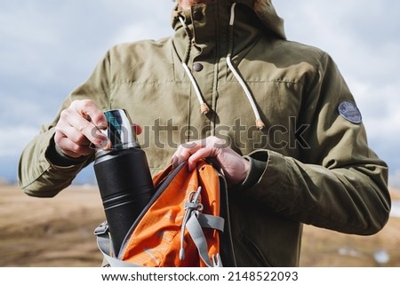 A tourist takes out from a backpack a black metal thermos, camping equipment, a vacuum thermos for hot drinks, equipment on a hike, dishes of a tourist. High quality photo