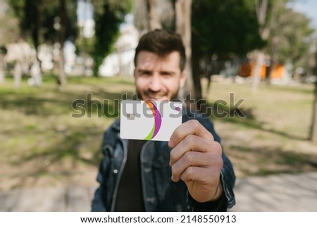 Young man wearing blue jean jacket holding a credit card to camera while standing on a city park. Happy man hands holding a debit card, focus on hands.