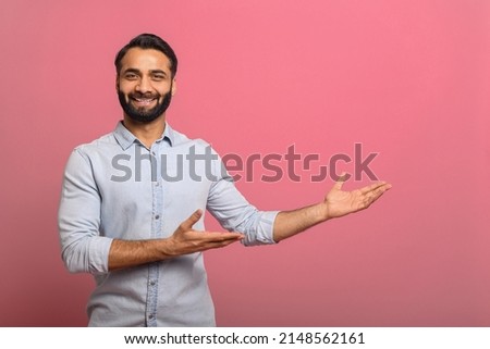 Handsome Indian guy points palms at empty copy space isolated on pink background. Cheerful multiracial bearded man in casual jeans shirt presenting novelty, advertising concept Royalty-Free Stock Photo #2148562161