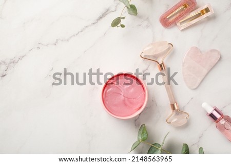 Skincare concept. Top view photo of two barrettes rose quartz roller gua sha pink eye patches dropper bottle with cosmetics and eucalyptus on white marble background with copyspace