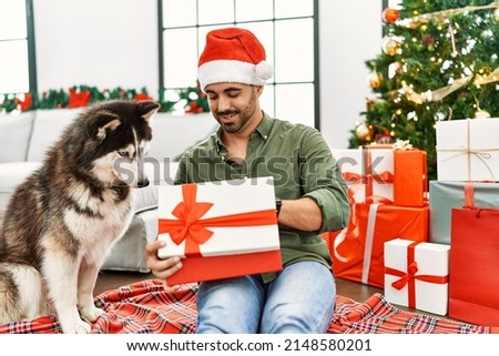 Young hispanic man unboxing gift sitting on floor with dog by christmas tree at home