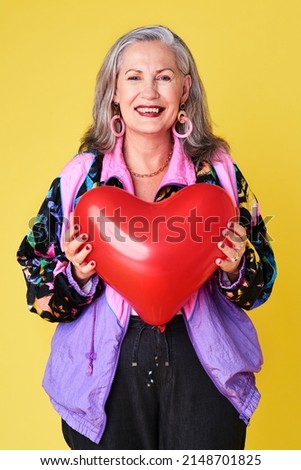 Always remember to spread love. Portrait of a confident and stylish senior woman holding a heart shaped balloon against a yellow background.