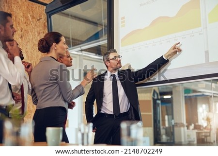 Running through the numbers with his team. Shot of an executive giving a presentation on a projection screen to a group of colleagues in a boardroom. Royalty-Free Stock Photo #2148702249