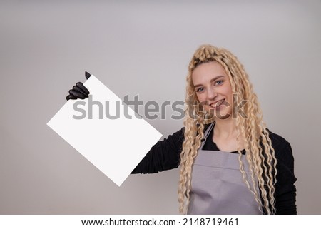 Happy european blonde girl with long hair holding a blank vertical canvas in her hands, poster mockup with empty frame. A specialist in the field of manicure, hairstyles, tattoo holds a certificate.