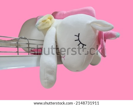 Cute white unicorn doll lying on iron railing on isolated pink background.
