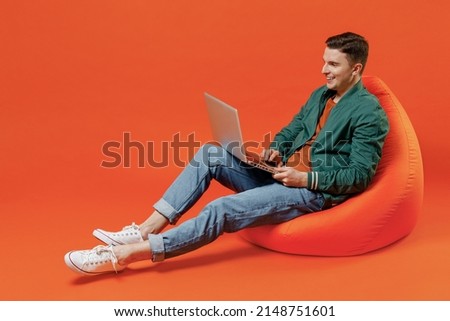 Full size body length happy smiling vivid young brunet man 20s wears red t-shirt green jacket sit in bag chair hold use work on laptop pc computer isolated on plain orange background studio portrait.