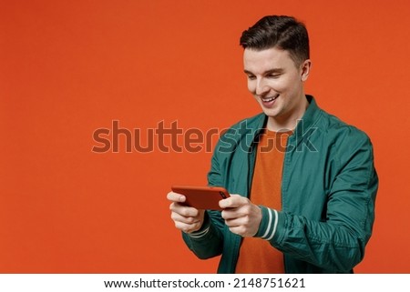 Gambling young brunet man 20s wears red t-shirt green jacket using play racing app on mobile cell phone hold gadget smartphone for pc video games isolated on plain orange background studio portrait