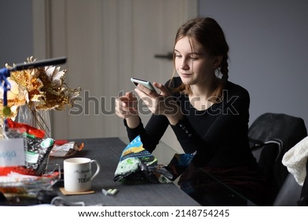 Girl in black T-shirt sits in kitchen holding phone in her hand and eating chips