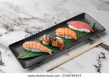 set of nigiri with tiger shrimp, tomago, salmon, tuna and green bamboo leaf in a black ceramic plate with chopstick on a bright white textured marble background, side view