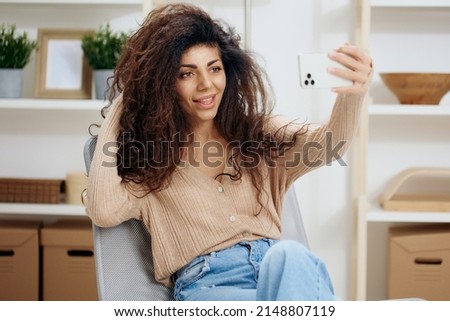Cheerful awesome tanned curly Latin lady taking selfie with trendy smartphone sit on chair in home modern interior look aside. Copy space Mockup Banner. Concept time to love yourself