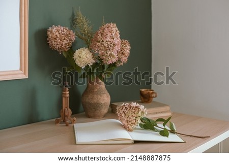 Aesthetic still life with hydrangea flowers, the spread of a book with blank pages, wooden candle holder, handmade ceramic cup on table near green and white wall.