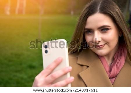 Young cheerful girl using smartphone over park background smiling looking to the phone and staring away thinking. Social network use by the phone.
