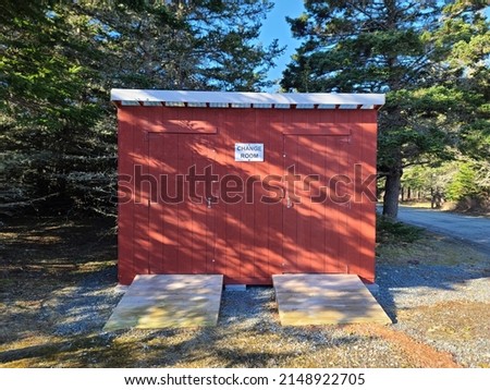 A small red building used as a changing room next to a forested area. There are two ramps leading to the separate rooms. It is handicap accessible.