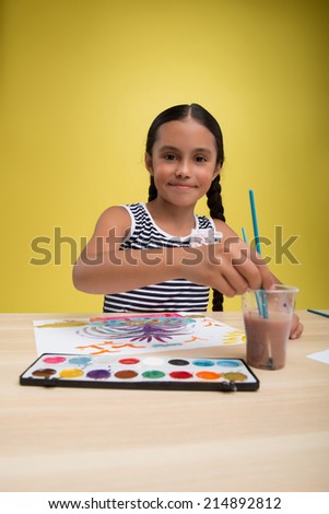 Half-length portrait of pretty dark-haired smiling little girl wearing nice striped dress with white rose on it sitting at the table soaking the brush in the water and painting her picture. Isolated