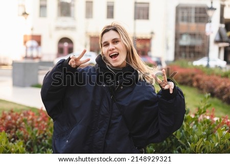 Cool blonde woman in navy blue bomber shows sign v, peace and friendship with two hands, open mouth and posing outside. Happy woman walk and posing outdoor in the city. 