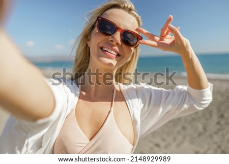 Lovely blonde female blogger wearing sunglasses showing peace sign, taking selfie on the beach