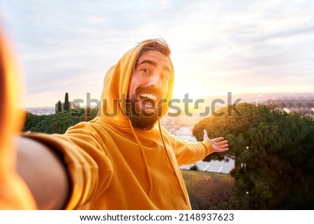 Selfie of young smiling man hooded sweatshirt. Happy Guy with good laugh taking self picture at sunset showing beautiful views. Cheerful traveler enjoying photographing their vacations having fun