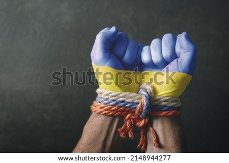 War in Ukraine. Male hands in the colors of the flag of Ukraine tied with a rope in the colors of the flag of Russia on a dark background. Conceptual image of violence, occupation, aggression Royalty-Free Stock Photo #2148944277
