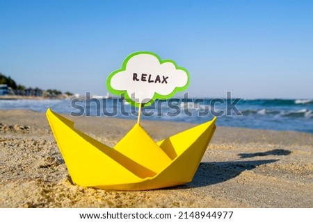 Small origami yellow paper boat with stick and speech bubble with word RELAX stands on sandy beach near sea water and sea waves with white foam close-up. Concept of travel, tourism, vacation, holiday