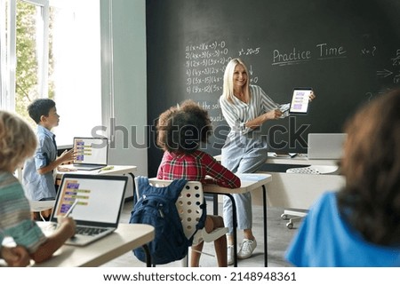 Smiling female teacher showing new maths task to elementary middle diverse schoolchildren sitting on desk at chalkboard holding tablet device. Kids using laptops. Education tech concept.