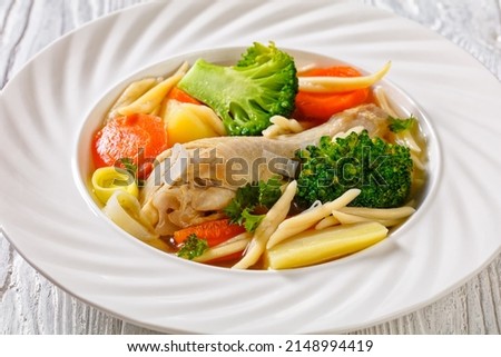 chicken vegetable soup with broccoli, carrots, parsnip, leek and pasta in white bowl on white wooden table, horizontal view from above, flat lay, free space