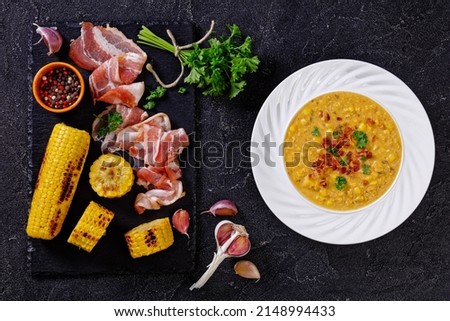 cheesy corn chowder topped with crumbled crispy fried bacon in white bowl on concrete table with ingredients, horizontal view from above, flat lay