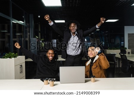Happy and joyful multiethnic men raise their hands and fists and rejoice in winning in online casino on laptop. Teambuilding of black Africans and Asian programmers