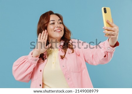 Young redhead chubby overweight woman 30s with curly hair wear pink shirt casual clothes doing selfie shot on mobile phone post photo on social network waving hand isolated on pastel blue background