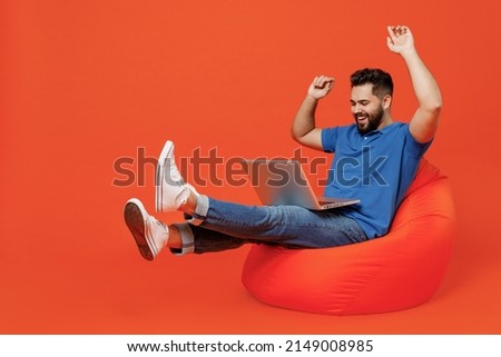 Full body young excited happy caucasian man wear basic blue t-shirt sit in bag chair hold use work on laptop pc computer isolated on plain orange background studio portrait. People lifestyle concept