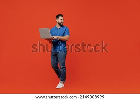 Full body young smiling happy caucasian man 20s wear basic blue t-shirt looking aside on workspace area hold use work on laptop pc computer isolated on plain orange background People lifestyle concept