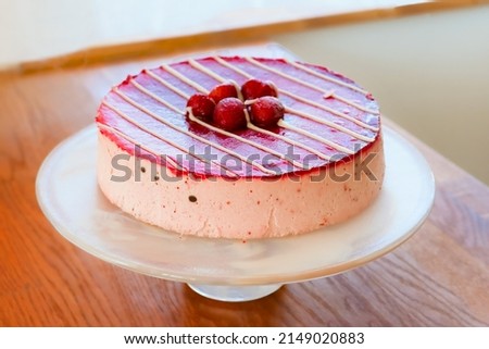ruit and chocolate cake of red-raspberry color in a white parallel strip of glaze with strawberries on a glass dish on a wooden tabletop by the window
