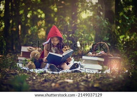 Books can transport us to the most magical places. Shot of a little girl in a red cape reading a book with her toys in the woods. Royalty-Free Stock Photo #2149037129