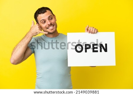 Brazilian man over isolated purple background holding a placard with text OPEN and doing phone gesture