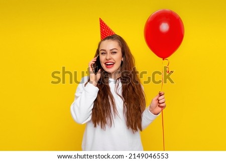 Positive young woman is talking on a mobile phone, receives congratulations from friends on anniversary, wears party cone and white casual style sweater, feels very happy, yellow background