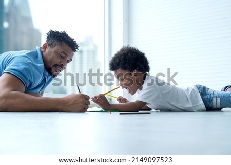 Happy African family, lying on the floor at home, father helping little cute curly son drawing picture, out of window high building view in blur background