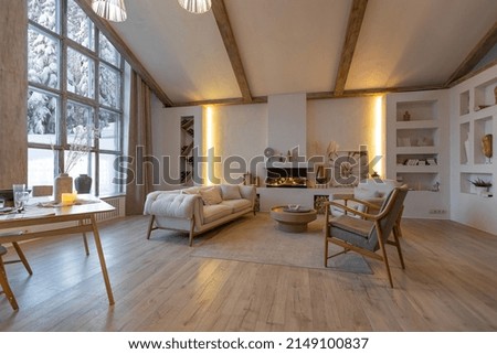 cozy warm home interior of a chic country chalet with a huge panoramic window overlooking the winter forest. open plan, wood decoration, warm colors and a family hearth. Royalty-Free Stock Photo #2149100837