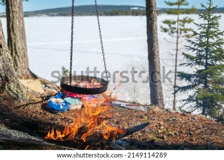 Frying pan full of  bacon hanging over a camp fire, frozen lake and blue sky in background.
Picture from Vasternorrland Sweden.
Focus on foreground.