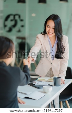 Two young Asian businesswomen join hands and work together as a team for success in business and finance, greetings and alliance ideas. with documents, graphs, and tablets on the table