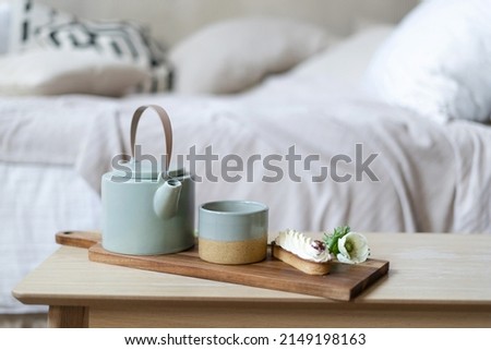 Still life details in home interior of bright bedroom in bohemian chic style. Close up cup of tea, teapot and eclair on serving tray stand at coffee table. Breakfast against blurred bed on background