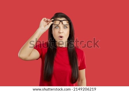 Beautiful young brunette woman impressed by something extraordinary looks at camera with surprised, shocked, astonished face expression, lifts her glasses and says WOW. Studio shot on red background