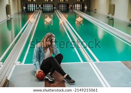 Young exhaustion woman sitting on a lane with bowling ball and feeling disappointed after failure throwing at the bowling club.